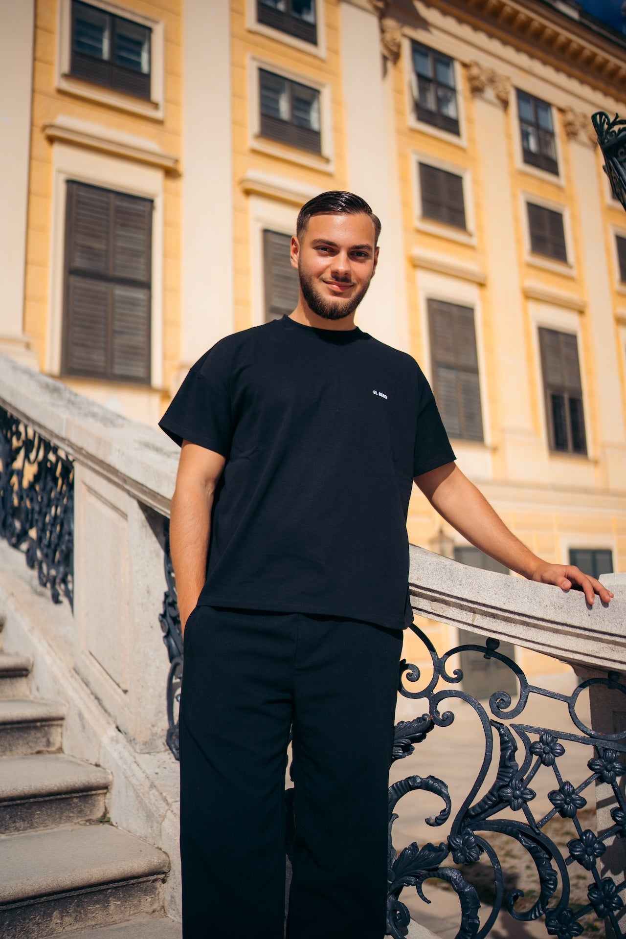 Man wearing a black t-shirt and pants standing on a balcony with a building in the background